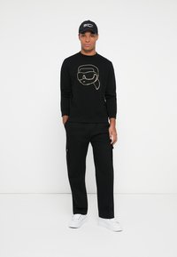 Felpa nera con un design grafico dorato di una persona che indossa occhiali protettivi, abbinata a pantaloni cargo neri e sneakers bianche. Cappellino nero in testa.