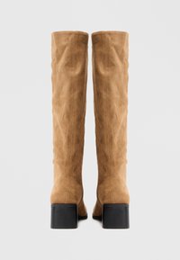 Paire de bottes en suède beige hautes avec des talons carrés, vues de l'arrière sur un fond clair uni.