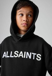 Chico con pelo oscuro vistiendo una sudadera negra con el texto "ALLSAINTS", mirando hacia un lado contra un fondo claro y sencillo.