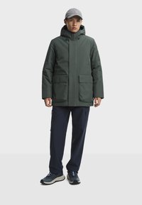 Zelená zateplená bunda s kapucňou, dvoma prednými vreckami, nosená cez námornícke cargo nohavice a tmavé topánky s modrými akcentmi, doplnená sivou čiapkou.