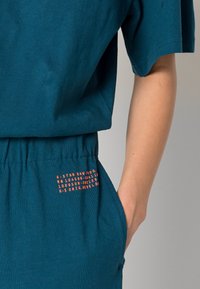 Turquoise katoenen top met korte mouwen, voorzien van een elastische tailleband en oranje tekst aan de zijkant. Vlotte textuur en ontspannen pasvorm.
