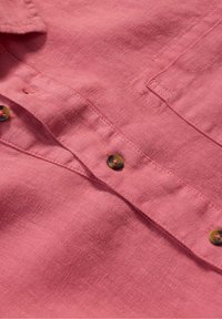 Rosa linneskjorta med en strukturerad yta, prydd med bruna knappar och en klassisk krage. Närbild på tyget och detaljer.