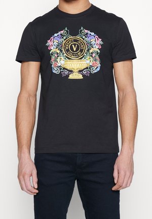 Man draagt zwart T-shirt met kleurrijk, sierlijk Versace Jeans Couture-logo en gouden trofee-ontwerp aan de voorkant.