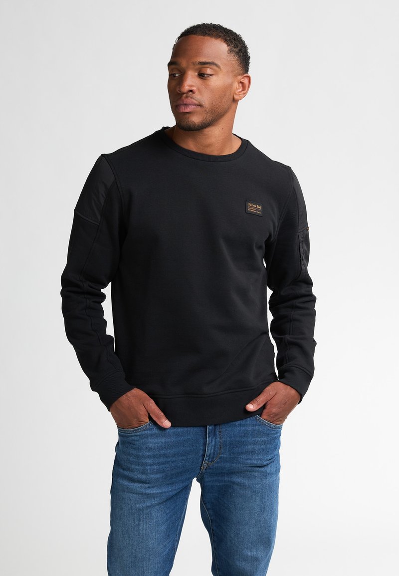 Zwart sweatshirt met lange mouwen, geribbelde manchetten, ronde hals, textuurstof en een klein merkembleem op de borst. Gecombineerd met blauwe jeans.