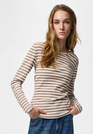 Fiatal nő hosszú szőke hajjal, bézs-fehér csíkos hosszú ujjú felsőt és kék farmert visel, kezét a zsebében, semleges kifejezés.