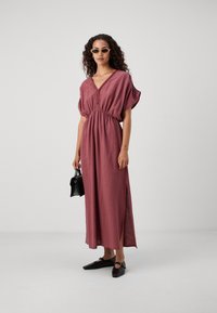 Robe maxi de couleur rose avec une silhouette décontractée, taille froncée, décolleté en V profond et manches courtes. Associée à des accessoires et des chaussures noires.