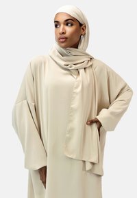 Beige, flydende caftan med brede ærmer og et draperet tørklæde. Glat stof med en subtil glans. Modellen poserer med den ene hånd på hoften.
