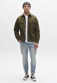 Cămașă kaki cu nasturi și buzunare aplicate, tricou alb, blugi albaștri deschiși cu manșete rulare și adidași negri cu talpă albă.