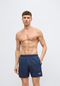 Tengerészkék fürdőnadrág elasztikus derékpánttal és oldalzsebekkel, könnyű anyagból készült. Az elején egy kis fehér logó található.