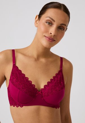 Donna con capelli scuri e trucco leggero che indossa un reggiseno di pizzo fucsia brillante con bordi smerlati, su uno sfondo chiaro uniforme.