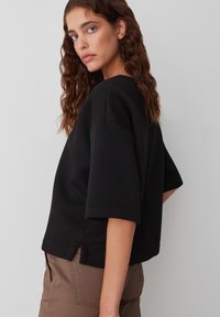 Felpa nera con maniche ampie, colletto a coste e spacchi laterali. Indossata con pantaloni beige, caratterizzati da un tessuto liscio e testurizzato.