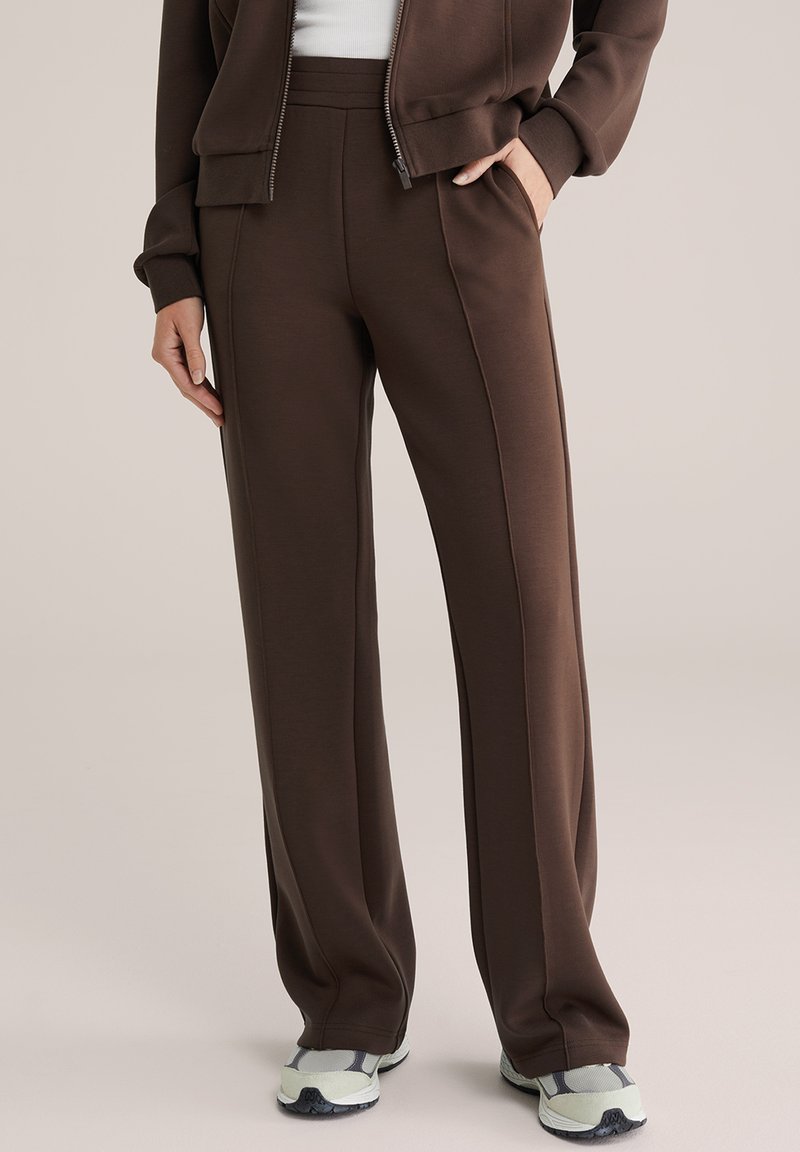 Ember barna egyenes szárú nadrágban és hozzáillő cipzáras dzsekiben, egyik kezével a zsebében áll, fehér tornacipő látható.