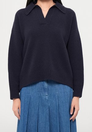 Mujer con un suéter de punto azul marino oscuro con cuello y escote en V, combinado con una falda plisada de denim azul.
