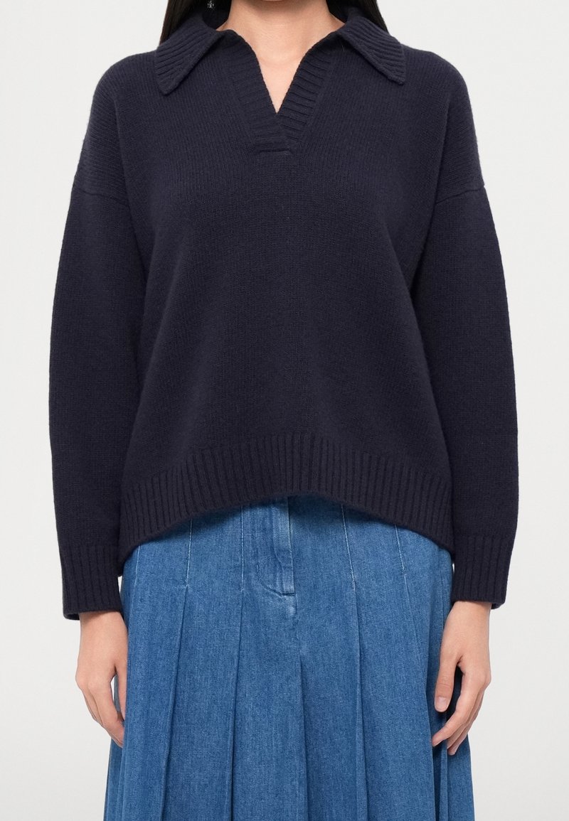 Femme portant un pull en tricot bleu marine foncé avec un col et un décolleté en V, associé à une jupe plissée en denim bleu.