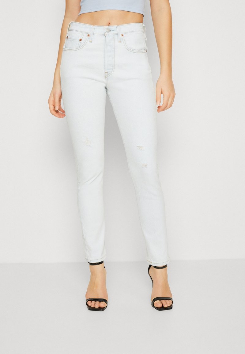 Jeans ajustados de cintura alta en un azul claro con detalles desgastados y estilo tradicional de cinco bolsillos, combinados con sandalias de tacón alto negras con tiras.