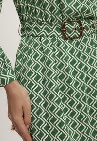 Zielona sukienka z geometrycznym białym wzorem, z taką samą paskiem podkreślającym talie oraz teksturowaną tkaniną ilustrującą fałdy.