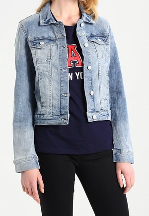 Femme portant une veste en jean bleu clair sur un t-shirt bleu marine avec texte rouge et blanc, associée à un pantalon noir, debout sur un fond blanc.