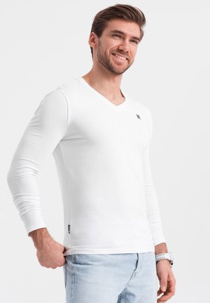 Camisa blanca de manga larga y escote en V con un corte ajustado, hecha de una suave mezcla de algodón. Detalle de pequeño logotipo cerca del pecho. Llevada con jeans de color azul claro.