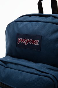 Blå Jansport ryggsekk med strukturert stoff, med et fremtredende logo-patch. Designet inkluderer en glidelås foran lomme og polstrede stropper.