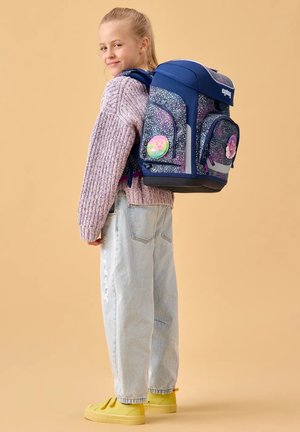 Girl wearing a purple sweater and light jeans with a large blue patterned backpack decorated with colorful pins, standing against beige background.