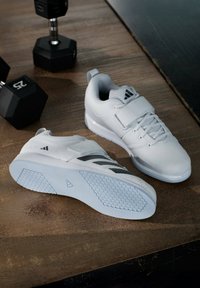 Białe buty sportowe z teksturowaną siateczkową cholewką, wspierającym paskiem i gumową podeszwą o sześciokątnym wzorze. Posiadają szare akcenty i oznakowanie.