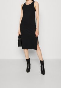 Robe noire sans manches avec encolure ronde et fentes sur les côtés, fabriquée en tissu extensible. Portée avec des bottines noires et une pochette.