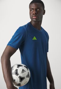 Navy blue athletic shirt with short sleeves and green Adidas logo, held with one arm, alongside a black-and-white soccer ball.