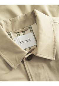 Bežna košulja s ovratnikom, koja se zatvara na gumb, s etiketom "LES DEUX." Unutra je vidljiv zeleni i krem prugasti uzorak.