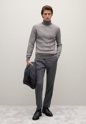 Pull à col roulé gris à côtes, associé à un pantalon gris, veste tenue à la main et bottines noires à cheville. Texture lisse, coupe moderne.