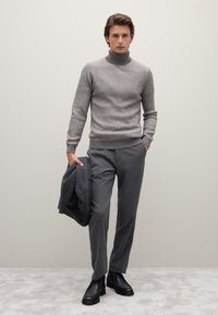 Grauer gerippter Rollkragenpullover kombiniert mit grauen Hosen, Jacke in der Hand gehalten und schwarzen Stiefeletten. Sanfte Textur, moderne Passform.