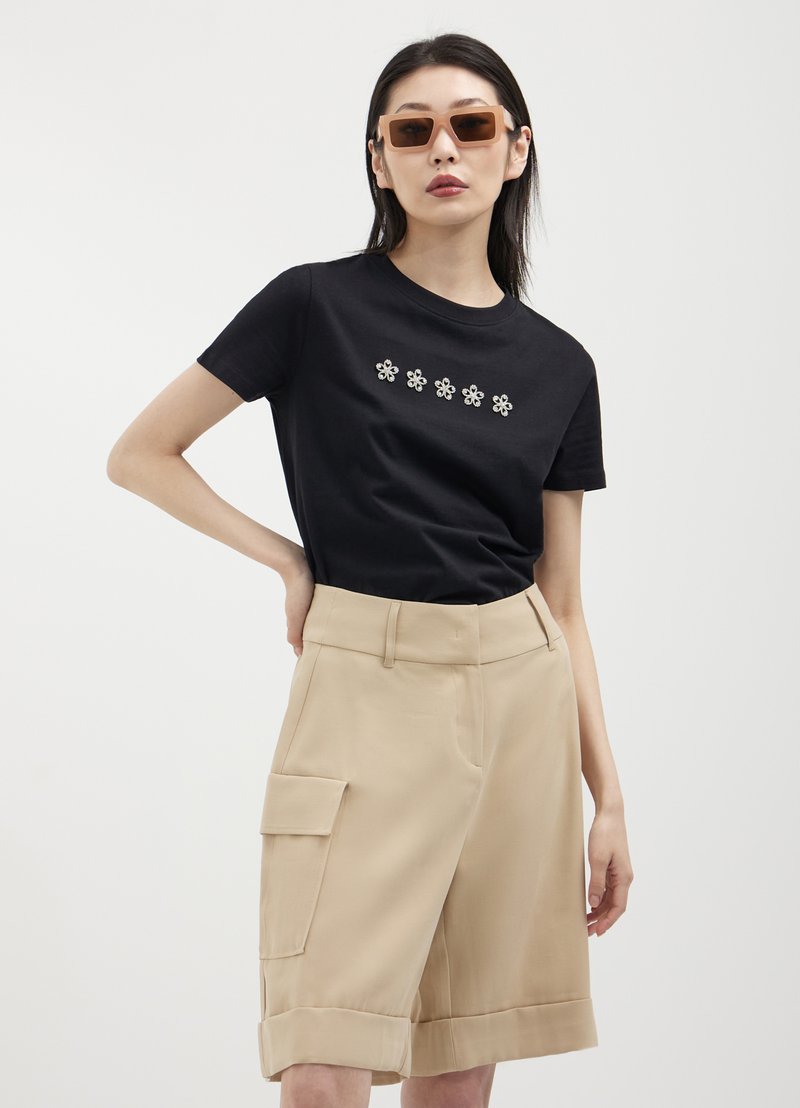 T-shirt en coton noir avec broderie florale, associé à un short cargo beige avec poches latérales et ourlet retroussé ; modèle portant des lunettes de soleil.