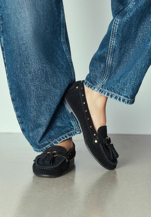 Persona che indossa jeans blu e mocassini in camoscio nero con nappine, in piedi su un pavimento grigio liscio.