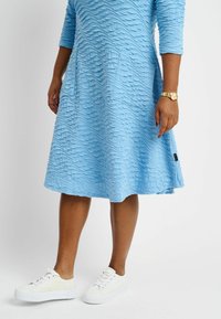 Helles blaues Kleid mit einem strukturierten, wellenförmigen Muster, Dreiviertelärmeln und einem ausgestellten Rock. Kombiniert mit weißen Sneakers und einer goldenen Armbanduhr.