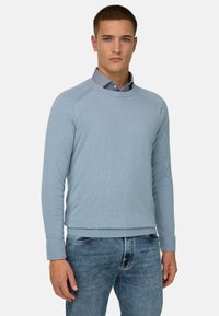Jeune homme portant un pull bleu clair par-dessus une chemise à col bleue et un jean bleu, debout devant un fond blanc uni.