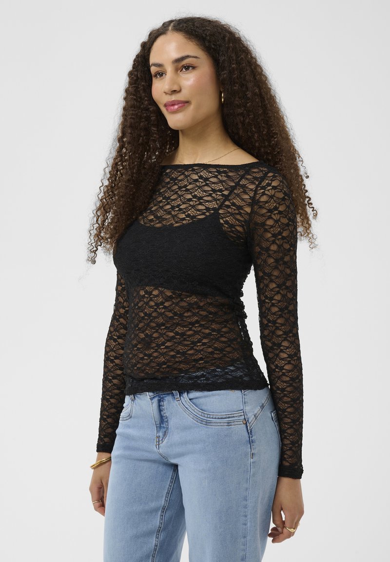 Femme aux cheveux bouclés portant un haut noir en dentelle à manches longues sur un caraco noir et un jean bleu clair, debout sur un fond uni.