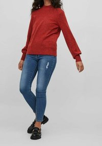 Czerwony sweter o fakturze, z okrągłym dekoltem, zestawiony z niebieskimi obcisłymi dżinsami i czarnymi trampkami z detalem w złotym kolorze.