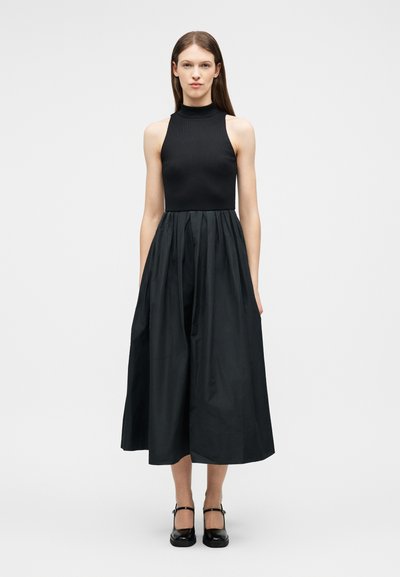 Femme debout portant une robe noire sans manches avec un col montant, un corsage ajusté et une jupe midi plissée et ample, associée à des chaussures Mary Jane noires.