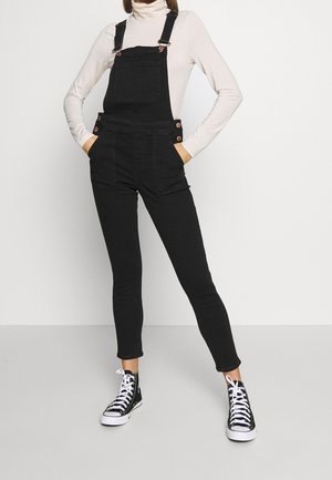 Persoon die zwarte denim tuinbroek, witte coltrui en zwarte hoge sneakers draagt, staand met handen in de zakken tegen een effen achtergrond.