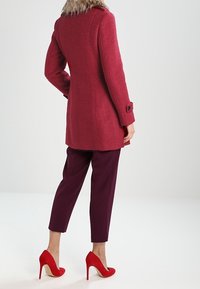 Punainen villakangastakki, jossa on turkiskaulus, mukautettu istuvuus ja nappihihnat, yhdistettynä syvän burgundin värisiin housuihin ja punaisiin korkokenkiin.