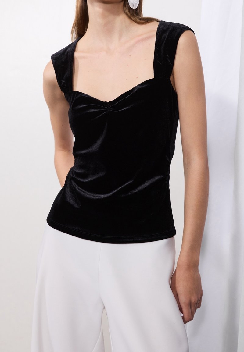 Top sin mangas de terciopelo negro con escote cuadrado, detalle fruncido en la parte delantera y tirantes anchos. Combinado con pantalones de pierna ancha blancos.