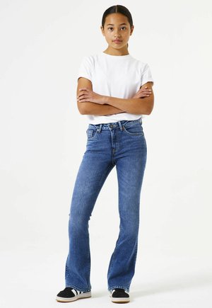 Blaue Jeans im ausgestellten Schnitt mit heller Waschung, fünf Taschen und einem Knopfverschluss. Kombiniert mit einem schlichten weißen T-Shirt mit kurzen Ärmeln und schwarzen Sneakers.