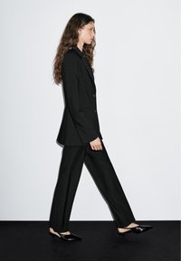 Zwart op maat gemaakt pak met een strak jasje en losse broek, gecombineerd met puntige zwarte schoenen. Glad textuur, minimalistisch ontwerp.