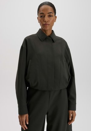 Donna con capelli scuri raccolti indossa una giacca nera larga con colletto e pantaloni neri abbinati, in piedi davanti a uno sfondo chiaro e uniforme.