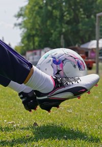 Bijele i crne nogometne kopačke s ružičastim naglascima i crnim vezicama, stopala lebde iznad šarenog nogometnog lopta na zelenoj travi.