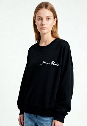 Noor naine sirgete blondide juustega kannab musta dressipluusi kirjaga "Mira Paris" ja siniseid teksapükse, seisab tavalise hele tausta ees.