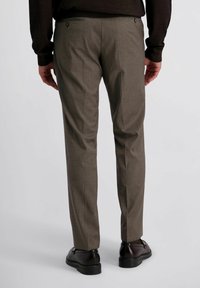 Man in een bruine broek, zwarte schoenen en een donkerbruin langetermijnshirt, staand met ontspannen handen langs de zij.