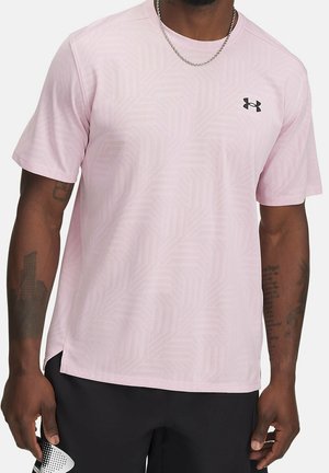 Mężczyzna w jasnoróżowej koszulce Under Armour z subtelnym geometrycznym wzorem i czarnych szortach, widoczne tatuaże na ramionach oraz srebrny łańcuszek na szyi.