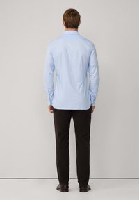 Camisa de manga larga azul claro con cuello, corte ajustado y puños con botones. Pantalones oscuros, zapatos elegantes, el fondo es liso y neutro.