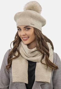 Béret en tricot beige avec un pompon en fourrure sur le dessus, associé à une écharpe assortie avec des bords côtelés, une texture douce et un design décontracté.