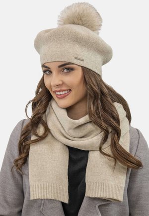 Béret en tricot beige avec un pompon en fourrure sur le dessus, associé à une écharpe assortie avec des bords côtelés, une texture douce et un design décontracté.